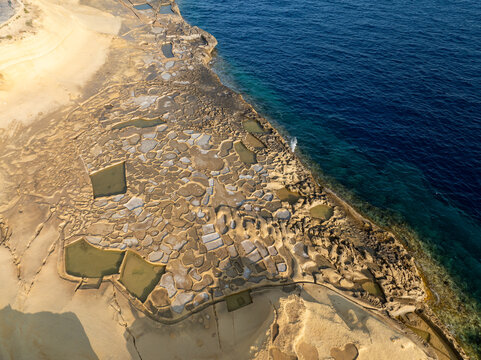 Aerial view of the ancient Qolla I-Bajda Salt Pans shimmer with golden light against the deep blue sea, creating a stunning tapestry of textures, Iz-Zebbug, Malta.
