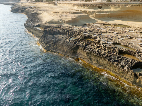 Aerial view of the rugged coastline meeting the turquoise sea near the Qolla I-Bajda Salt Pans, where the rocks are bathed in warm sunlight, Iz-Zebbug, Gozo, Malta.