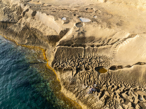 Aerial view of rugged, sun-baked rocks meeting the turquoise sea at the Qolla I-Bajda Salt Pans, Iz-Zebbug, Gozo, Malta.