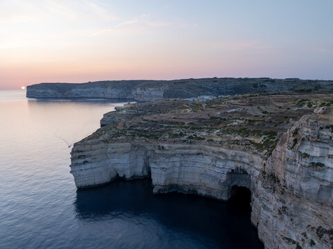Aerial view of rugged cliffs meet the tranquil blue sea under the soft glow of the setting sun at Sanap Cliffs, Munxar, Gozo, Malta.