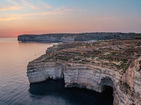 Aerial view of rugged cliffs meet the tranquil sea under a soft, pastel sky, where warm light kisses the horizon, Munxar, Gozo, Malta.