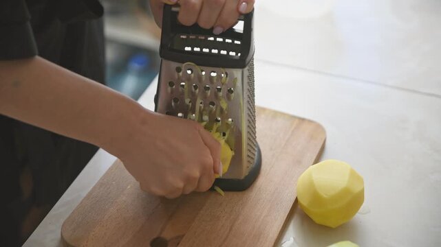 Hands grating peeled potatoes on metal box grater with wooden board background