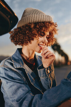 Curly woman wearing beige beanie and denim jacket smiles outdoors during sunset, enjoying a candid moment with soft natural light and blurred background, casual lifestyle vibe