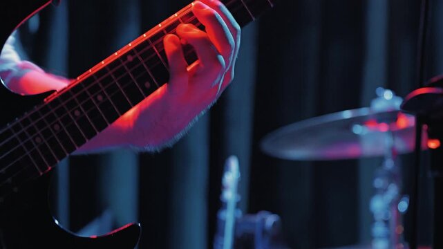 Close-up of a guitarist's hand on a fretboard playing fast. Dramatic purple stage lighting creates an energetic mood. Focus on the musician, with a blurred drum kit in the background