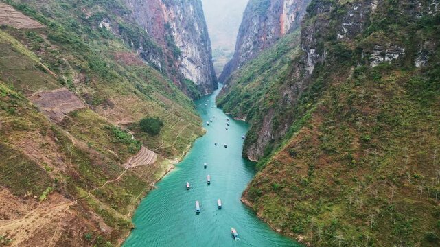 Aerial drone view of Tu San Canyon with a turquoise river winding through towering limestone cliffs and small boats drifting below in the rugged mountains of northern Vietnam
