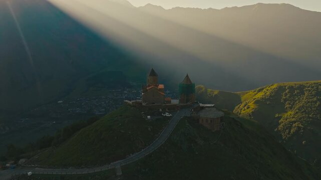 Drone shot of Gergeti Trinity Church in beautiful morning light with sun rays over Caucasus mountain peaks in Georgia, showing the historic site under renovation in a stunning alpine setting