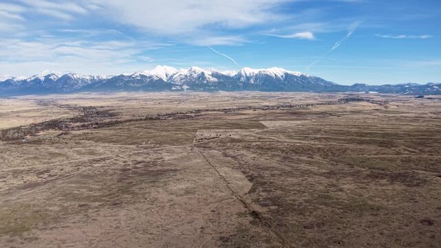 Aerial drone footage flying over a wide, open plain near Whitefish, Montana, reveals a dry prairie with sparse vegetation and snowcapped Rocky Mountains on the horizon under a bright sky.