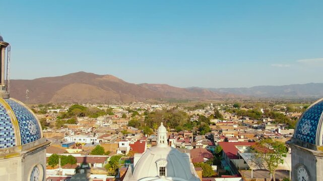 Drone footage captures the stunning architecture of Parroquia de San Juan Bautista in Tuxpan, Jalisco, under bright daylight. The video showcases the church's blue domes and the surrounding landscape.