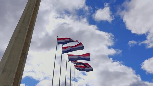 Flags flutter against a vivid blue sky in Plaza de Armas, Encarnacion. The landmark poles stand tall, capturing the serene yet vibrant atmosphere. Static, wide-angle shot.