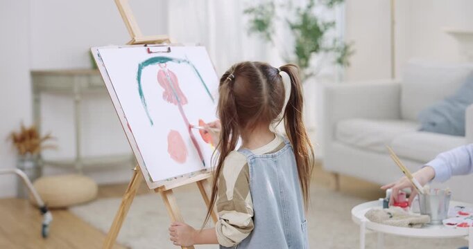 Creative playtime in living room as preschool daughter paints shape on easel canvas, Asian mother nearby preparing brush and color, art education for preschool child, imagination, bonding, home.