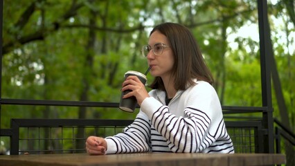 Fototapeta premium Young woman in a white sweater sipping coffee on a cafe veranda by the forest