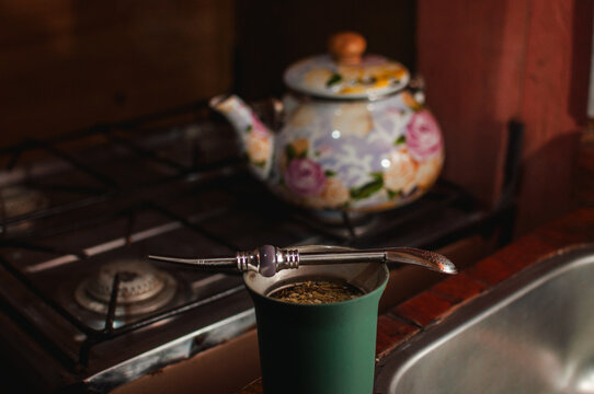 mate con yerba mate color verde sobre la mesa, tetera o pava sobre la cocina de fondo