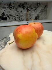 Obraz premium Two apples with red and yellow hues rest on a round, creamy marble surface. The background features a modern kitchen with a gold faucet and a black-white backsplash