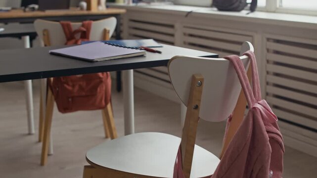 Wide establishing tilt up shot of empty modern classroom with notebooks on desks, backpacks hanging on chairs and chalkboard with economics theory