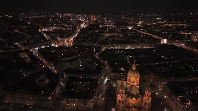 Stunning night vista of Munich from a drone. Highlighting the historic Frauenkirche cathedral towers and Marienplatz landmarks far behind the illuminated St. Lukas Church on the Isar banks