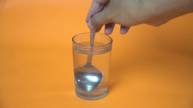 Close-up of a hand stirring clear water with a metal spoon inside a transparent glass