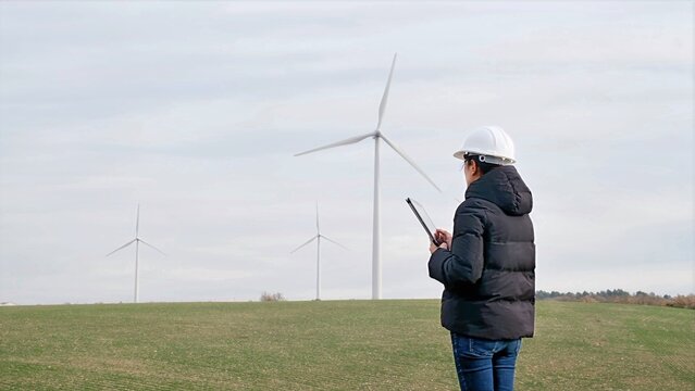 Female engineer in hard hat using tablet, inspecting wind turbines at a wind farm, managing green energy production and sustainable power generation outdoors