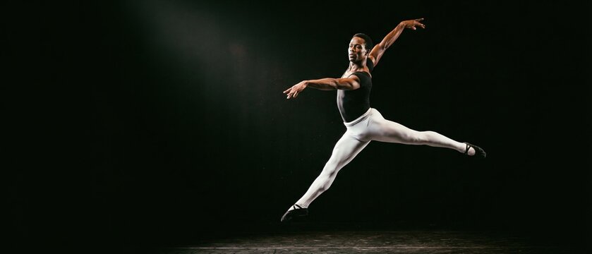 Male ballet dancer performing dramatic leap on dark stage with strong contrast lighting. Concept of theatrical performance scene, spotlight focus intensity, emotional expression through movement.