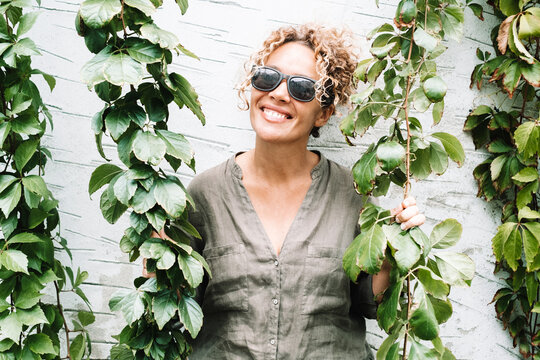 Smiling woman in sunglasses framed by green vines against a white wall enjoying outdoor relaxation