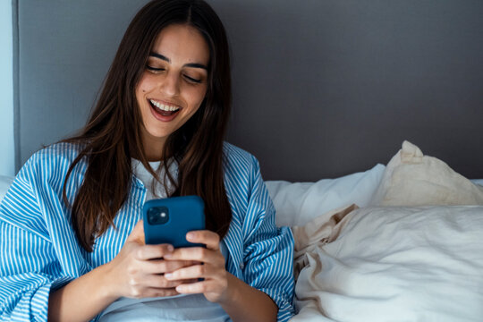 Smiling woman in striped shirt using a smartphone in bed, relaxing and messaging at home