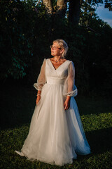 Fototapeta premium Female model wearing a white wedding dress with sheer sleeves stands in a lush green outdoor setting, sunlight filtering through trees in the background