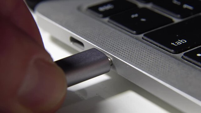 Extreme close-up of a hand plugging a grey USB Type-C cable into a modern silver laptop power port. Clean white background, high-end tech aesthetic.