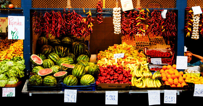 A market stall displaying various fruits and vegetables, including watermelons, peppers, bananas, oranges, and garlic.