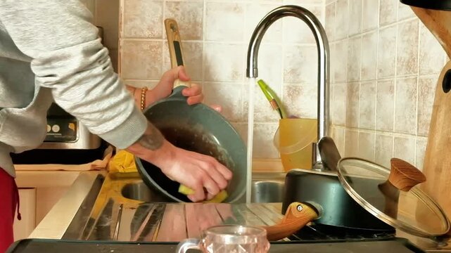 A man, whose arms are only framed, cleans a pan by rubbing it with a sponge. The man uses the kitchen sink as a support point
