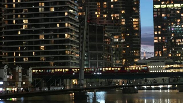 Night view in Canary Wharf, London capturing a DLR train moving across a bridge framed by illuminated skyscrapers. The scene highlights dense urban architecture, glowing windows, and reflections