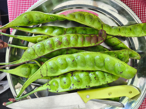 A silver plate filled with fresh petai pods and a knife on a red tablecloth, showcasing a traditional Indonesian ingredient.