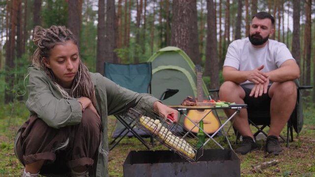 Young woman with dreadlocks kindling a fire in a brazier while her freelancer boyfriend works on a laptop at their campsite in a pine forest, enjoying an alternative lifestyle