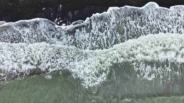 Dark rocks sit along the coast as strong waves roll in and crash against them. The water is white from foam and the sky above is gray with clouds.