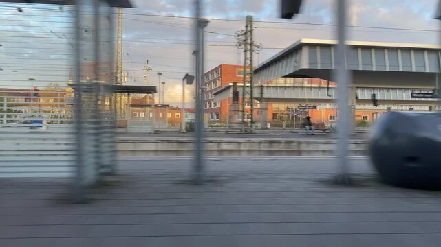 Point of view shot from a moving train window departing from Munster central station, passing platforms and urban buildings under a cloudy sky in Germany.