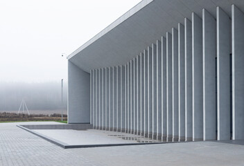 Fototapeta premium Curved colonnade of modern building stands beside empty paved space and open landscape. Repeating vertical columns create smooth architectural rhythm along exterior. Concept of public space, minimal