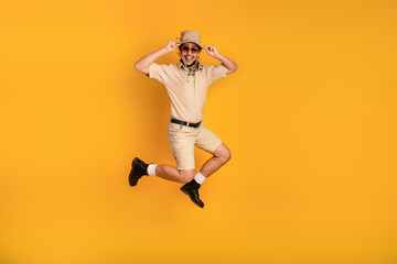 Obraz premium Happy young man in safari style outfit jumps mid air against bright yellow background showcasing adventure travel and summer fun