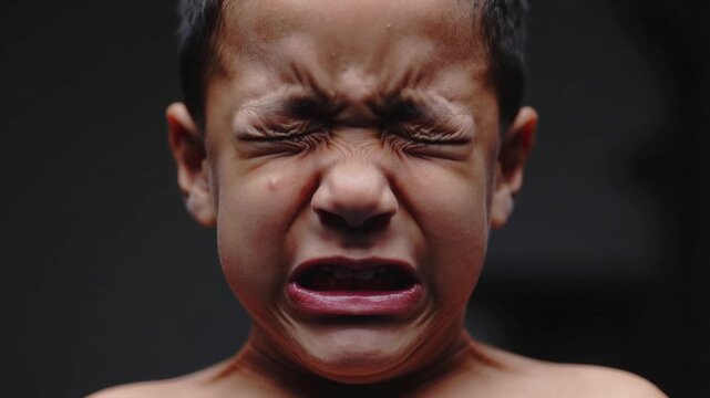 Filming shirtless child tightening facial muscles at studio dark background, tear showing emotion