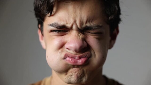 Puffing teen male with camera starting, pulling mouth and squinting eye in studio, brown shirt