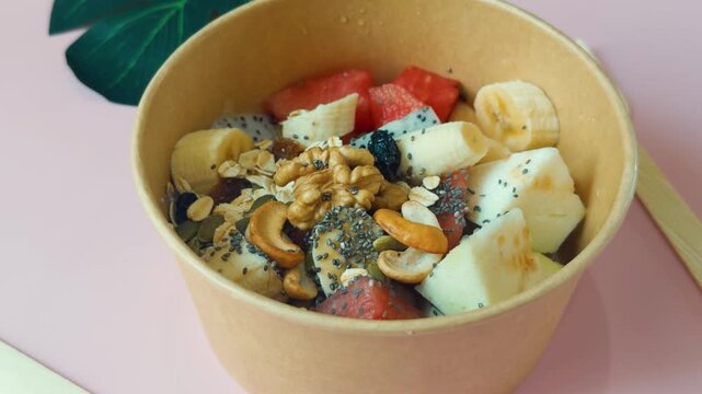 Customizing a healthy breakfast bowl with fresh fruit slices, crunchy granola, chia seeds, walnuts, and cashews, offering a nutritious and energizing meal on a pink background