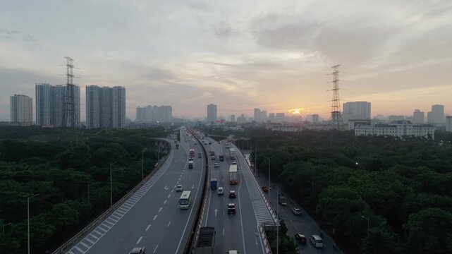 Drone perspective of multilane highway curving through green corridor in Hanoi during sunrise with vehicles moving steadily and distant high rise buildings