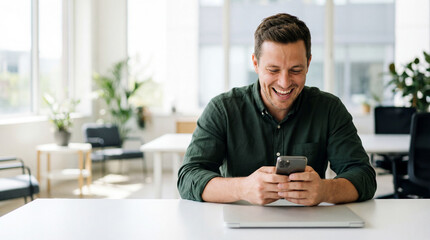 Man smiling at smartphone, happy man using phone at office desk, casual businessman laughing while texting, modern workspace natural light, close up shot
