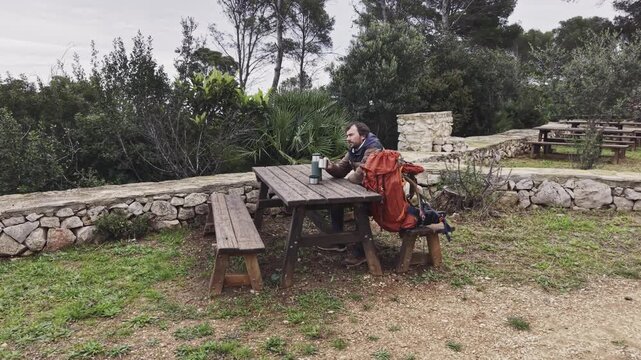 Man resting at picnic table drinking from mug during hiking trip