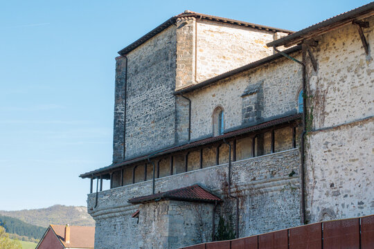 Fachada lateral de una iglesia medieval fortificada de piedra con galer&iacute;a porticada de madera y tejados de teja roja, bajo un cielo azul despejado en un entorno rural hist&oacute;rico.