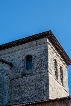 Fachada lateral de una iglesia medieval fortificada de piedra con galer&iacute;a porticada de madera y tejados de teja roja, bajo un cielo azul despejado en un entorno rural hist&oacute;rico.