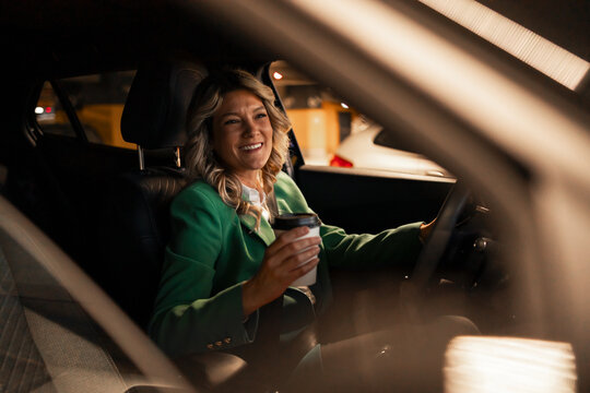 Businesswoman driving luxury car at night, smiling and drinking coffee