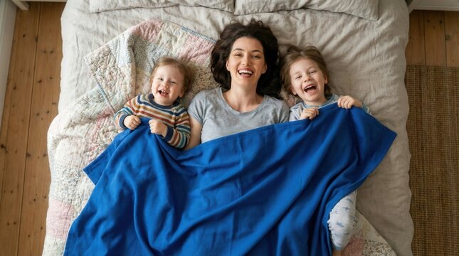 Joyful supermom with two cheerful children under a cozy blue blanket, lying on a bed with soft pastel linens, capturing a warm family moment from above