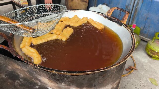 A large metal pot of frying oil with pieces of cakwe being deep-fried in a casual outdoor setting in Indonesia.