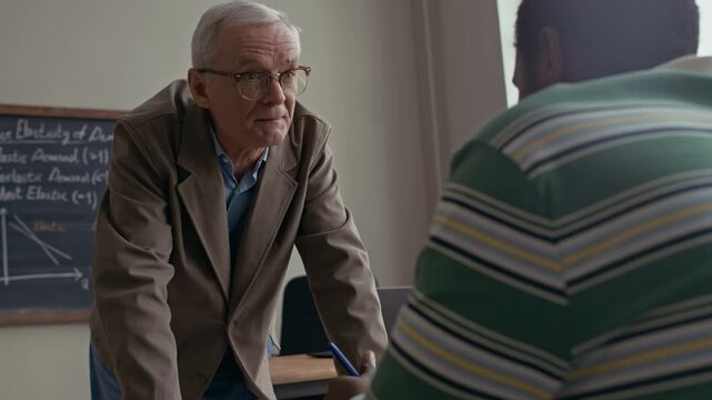 Over shoulder medium shot of senior Caucasian male teacher bending over desk helping African American student in bright classroom during lesson