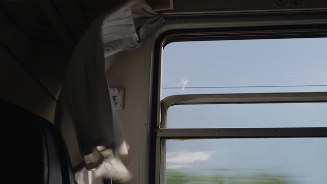 Interior view of a moving train compartment with a curtain fluttering in the wind. The landscape outside the window is blurred by speed