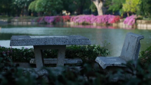 Stone concrete terrazzo table and bench set in a public park beside a green lake with pink bougainvillea flowers in bokeh