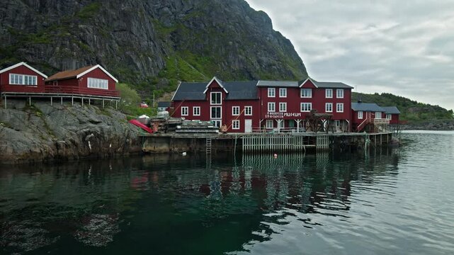 Traditional red wooden stockfish museum building on stilts above calm water in Lofoten, Norway. Scenic fishing village with mountain backdrop, coastal reflections, and Nordic maritime heritage.